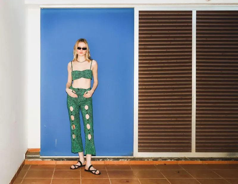 Model in a green croco-skin print bandeau top and cropped trousers with oval cut-outs, against a blue wall beside brown shutters