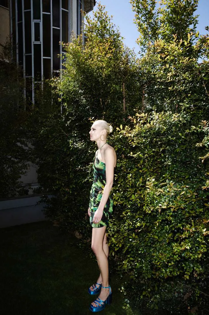 Model in profile in a green and black printed ruched mini dress and blue platform sandals beside a hedge