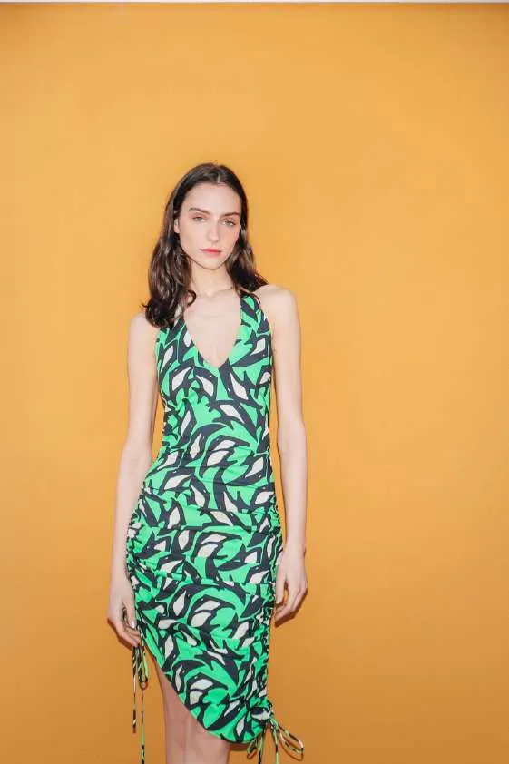 Model in a green, black and cream leaf-shape print halter dress with ruched side ties, against an orange wall