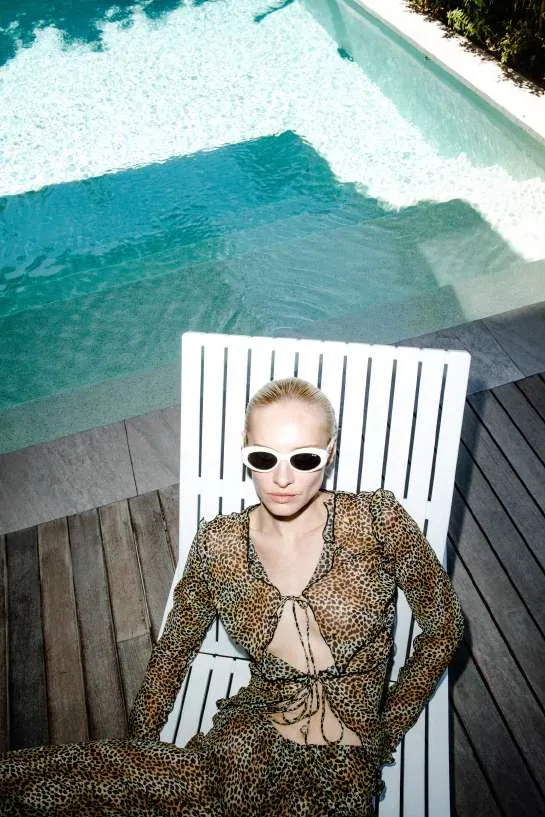 Model in white sunglasses and a sheer leopard-print tie-front top and trousers on a white lounger by a pool