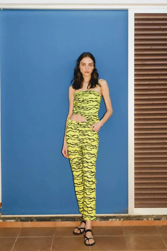 Model in a yellow and black wave-print bandeau top and trousers, against a blue wall beside brown shutters