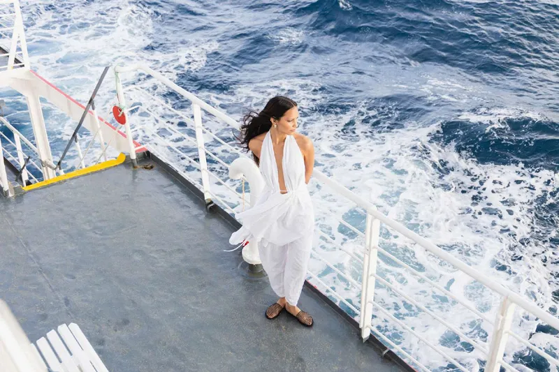 Model in a white halter top and draped trousers on the deck of a ferry, sea wake behind