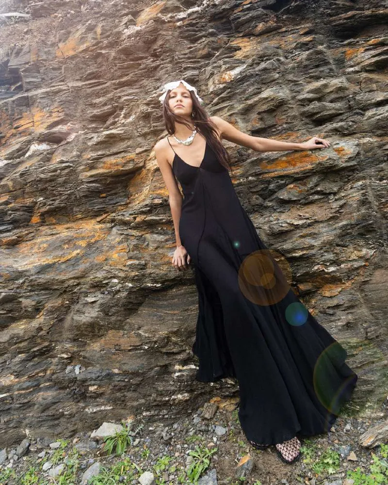 Model in a long black slip dress and a white headscarf, leaning against a layered rock face