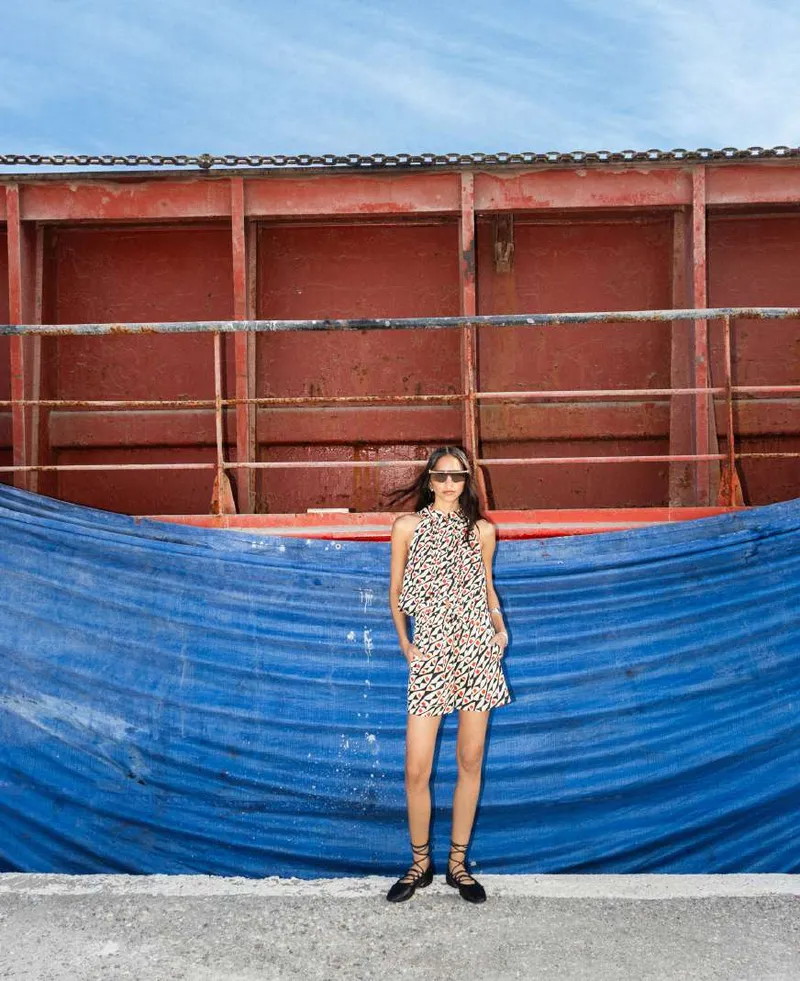 Model in a black, white and coral print halter mini dress in front of a blue tarpaulin and a rusted red container