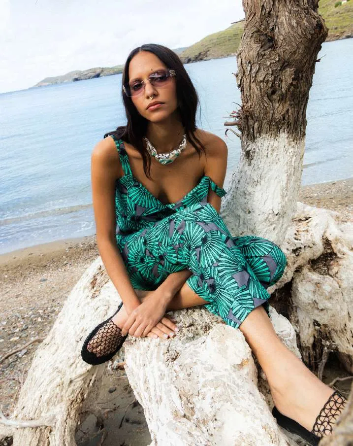 Model in a green and grey palm-leaf print slip jumpsuit, seated on a whitewashed tree trunk on a beach