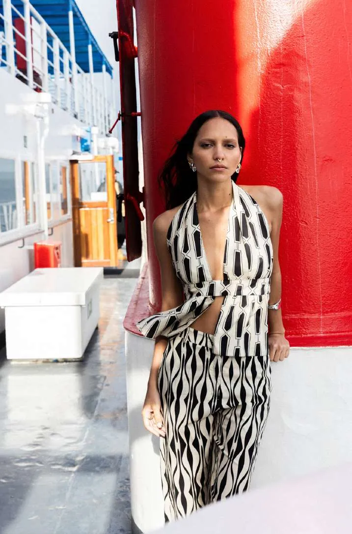 Model in a black and white wave-stripe halter top tied at the front and matching wide trousers, beside a red pillar on a ship's deck