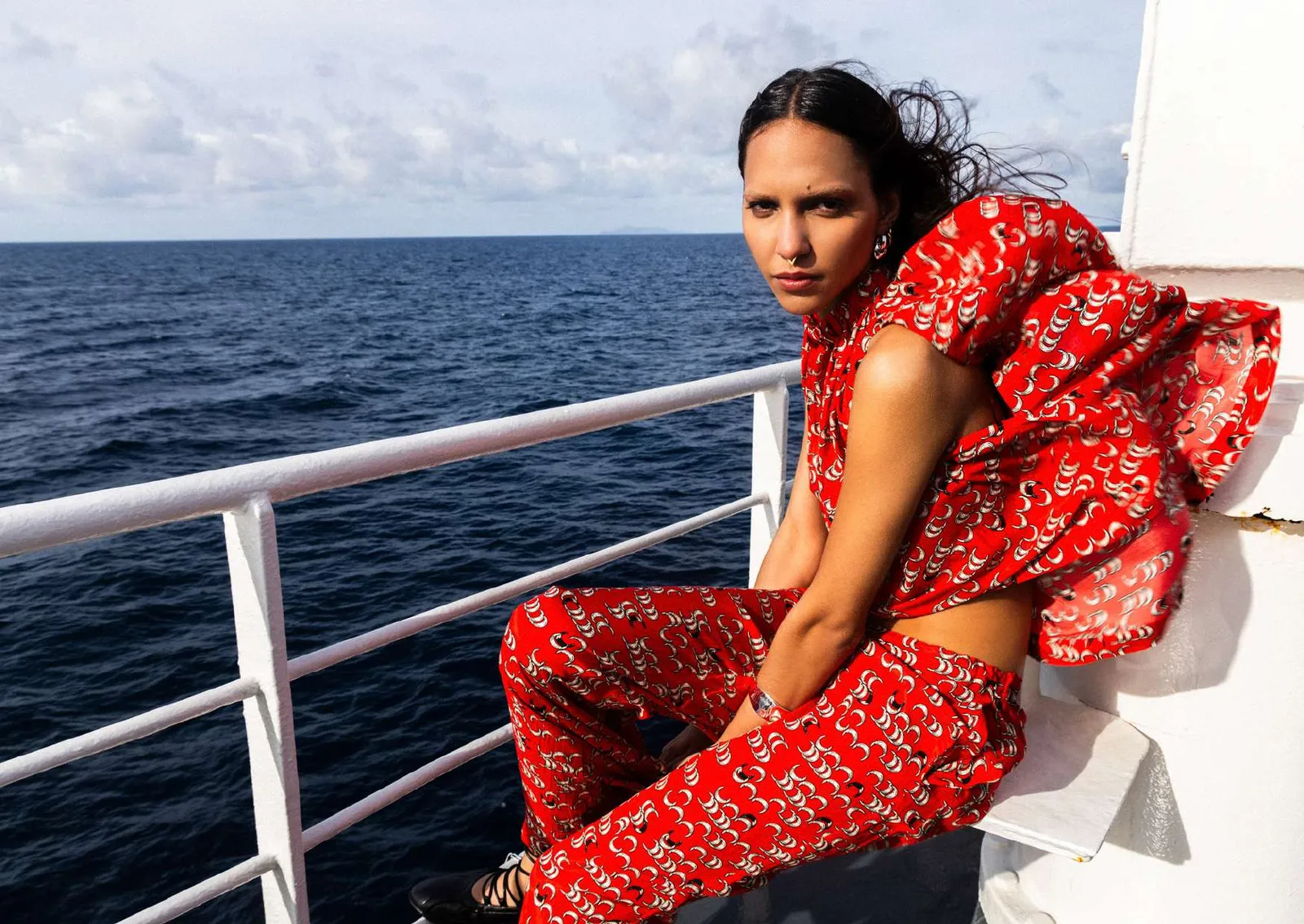 Model in a red and white crescent-print ruffled top and matching trousers, seated on a ferry railing with the open sea behind