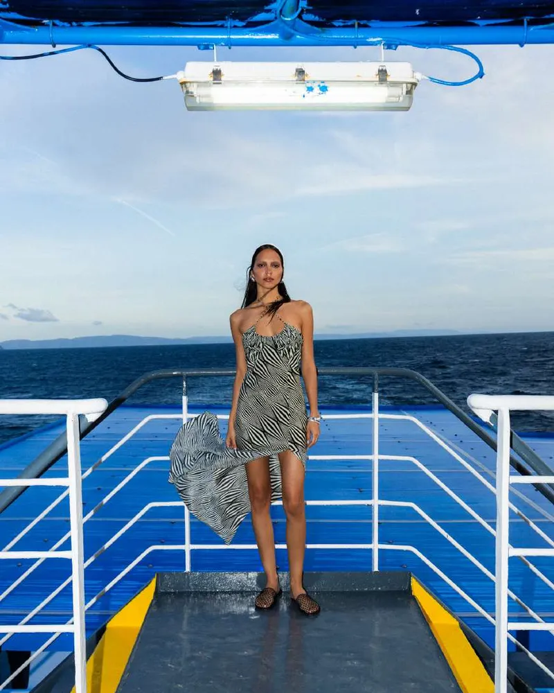 Model in a black and white zebra-stripe strapless dress with a long asymmetric hem lifted by the wind, on a ferry bow with blue decking