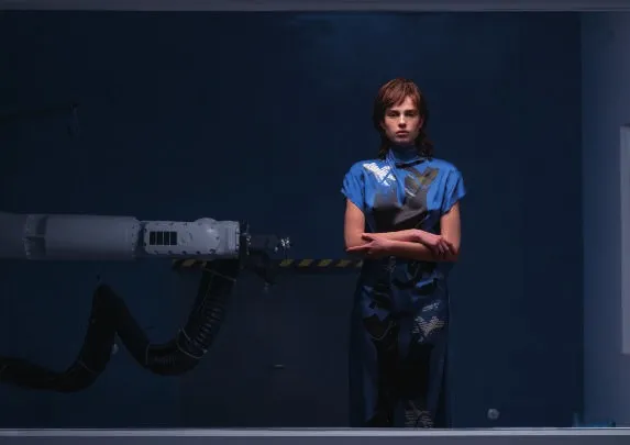 Model in the No Signal dress, blue printed silk, arms folded, beside a robotic arm in a dark blue room