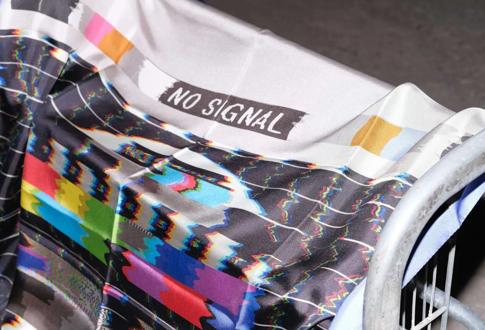 Close-up of the No Signal scarf, test-card colour bars broken by glitch lines, over a metal trolley