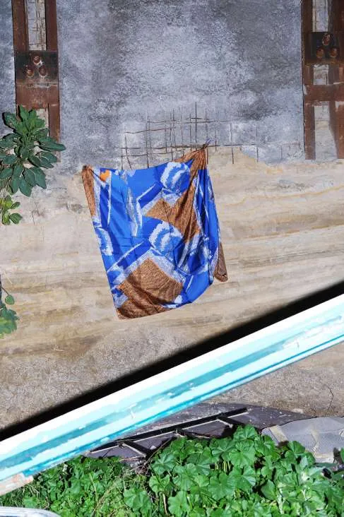 The Other Side scarf, blue and copper, hung on rusted rebar against a concrete wall