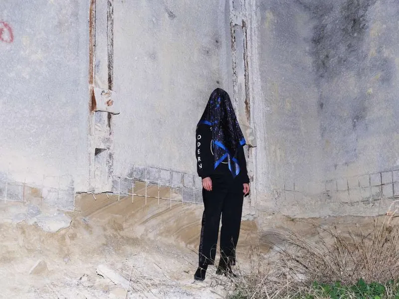 A figure in black with a navy printed scarf over the head, in front of a pale concrete wall