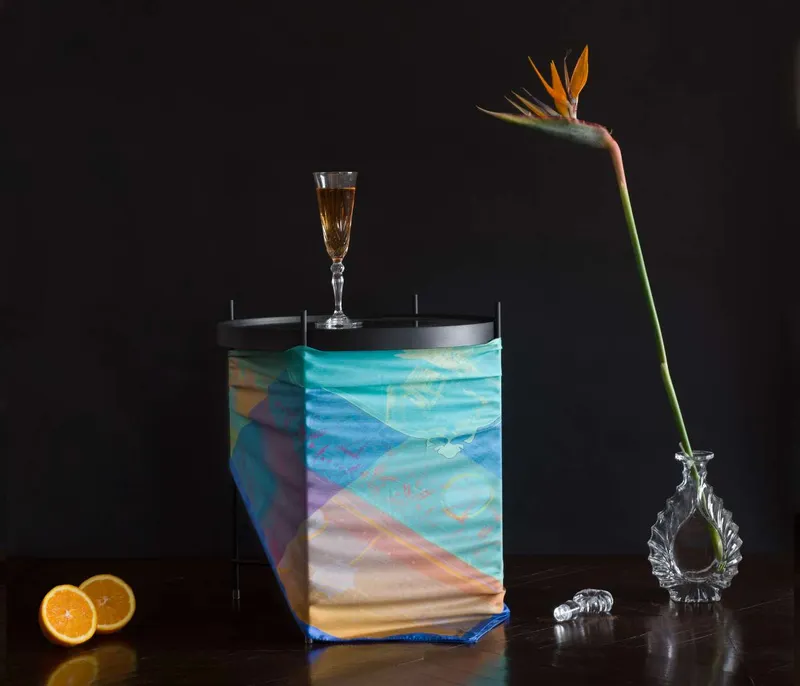 Still life on black: the Harmony scarf, blue, turquoise and pink, wrapped around a round side table with a glass of champagne on top, a halved orange and a bird of paradise in a glass vase