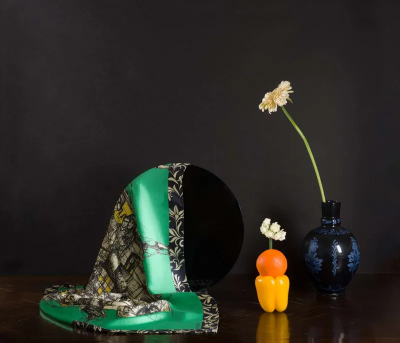 Still life on black: the Kalamos scarf, green with an ornate pattern, draped over a round black mirror beside an orange, a yellow pepper and a navy vase with a single flower