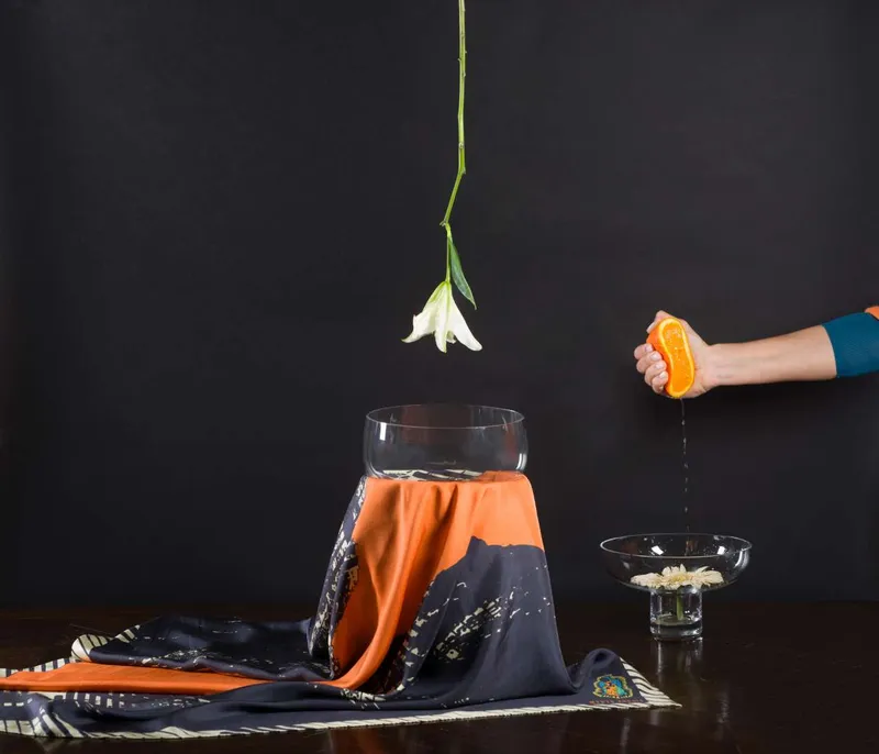 Still life on black: the Loupe scarf, orange and black, under a glass jar, a white lily hanging above and a hand squeezing an orange into a glass bowl