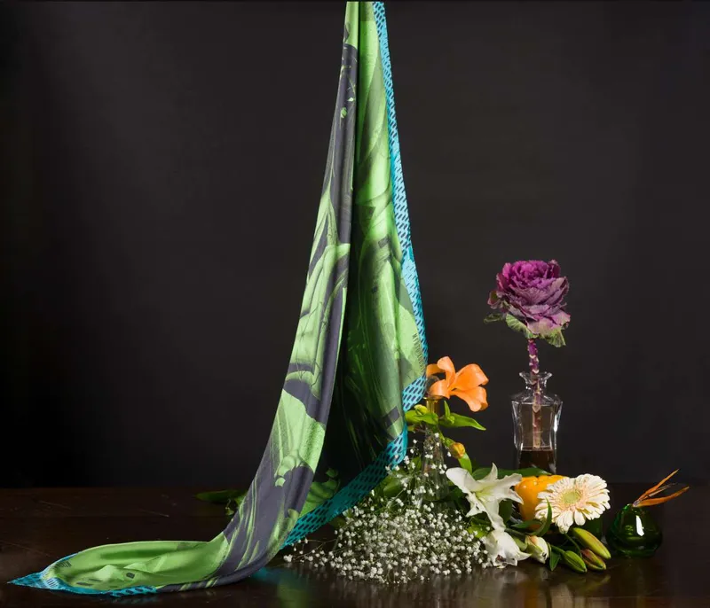 Still life on black: the Tires and Crosses scarf, green and teal, falling from above over a table with a purple rose, lilies, a gerbera and gypsophila