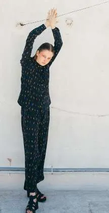 Model in a black shirt and trousers with a multicoloured dash print, arms raised, against a white wall