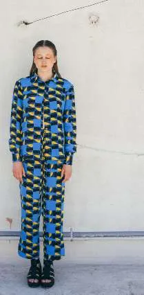 Model in a blue, black and yellow block-print shirt and trousers, against a white wall