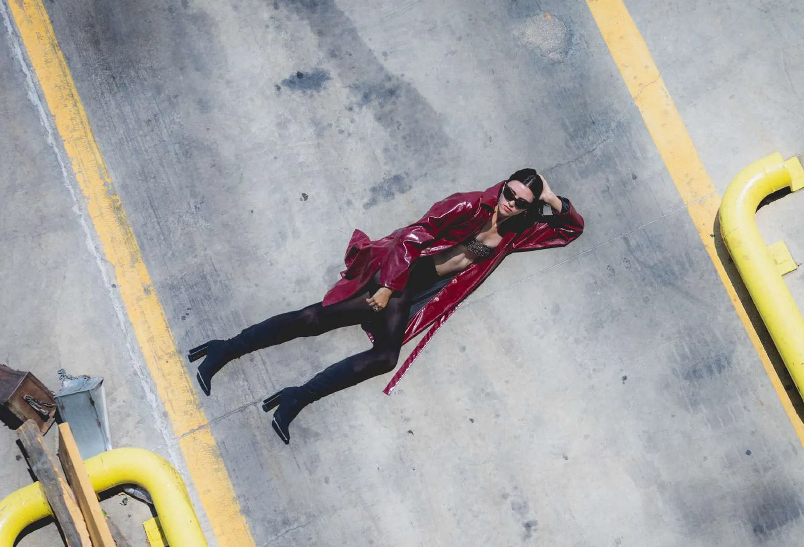 Model in a burgundy patent trench coat lying on a concrete ramp between yellow lines, seen from above