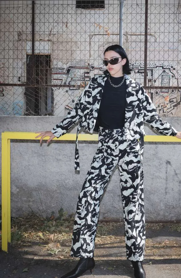 Model in a black and white abstract-print jacket and trousers over a black top, in front of a chain-link fence and a yellow rail