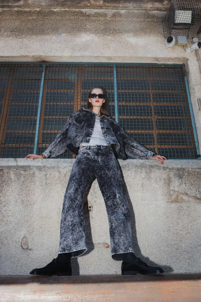 Model in a grey acid-wash denim jacket over a white top and wide acid-wash jeans, seen from below on a concrete ledge