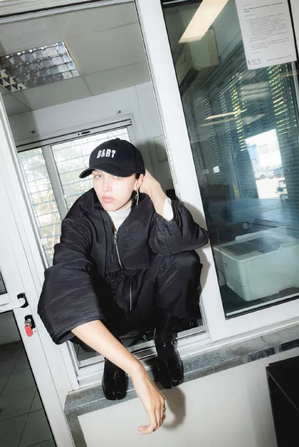 Model in an oversized black zip jacket, black trousers and a black cap, crouched on a window ledge in an office