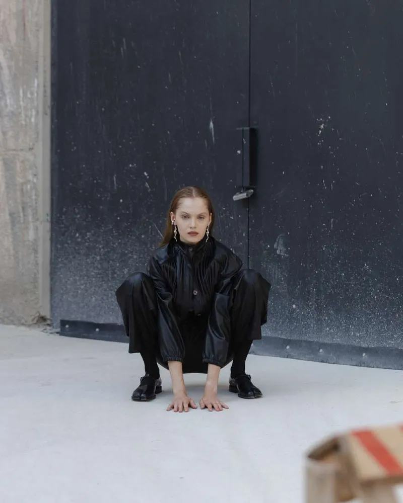 Model in a black leather-look shirt and trousers, crouching in front of a dark steel door