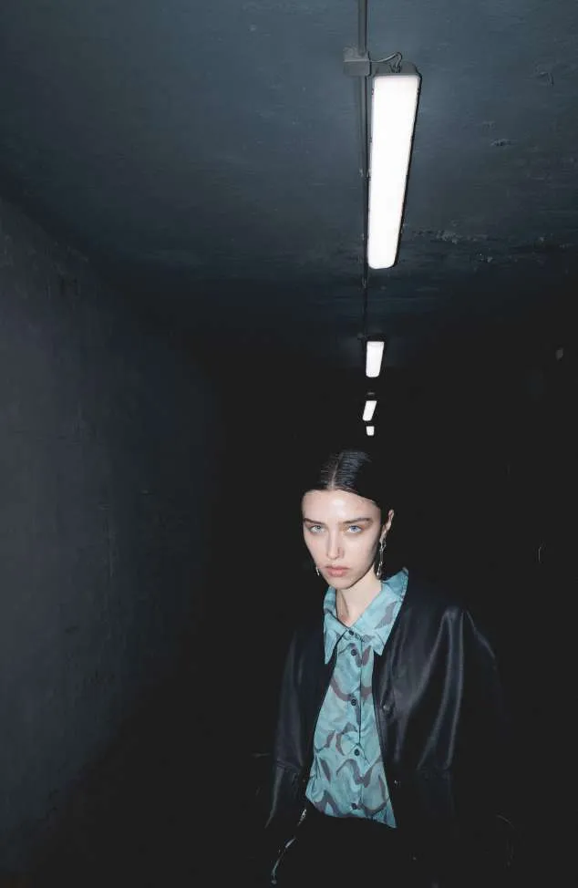 Model in a black leather-look jacket over a pale blue printed shirt, in a dark corridor under strip lights