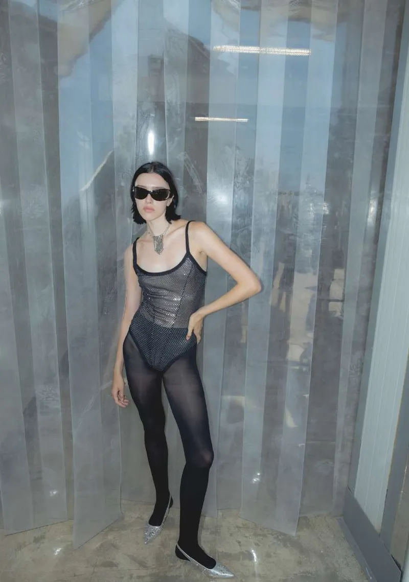 Model in a sheer black mesh bodysuit, black tights and silver pointed shoes, standing among translucent plastic curtains
