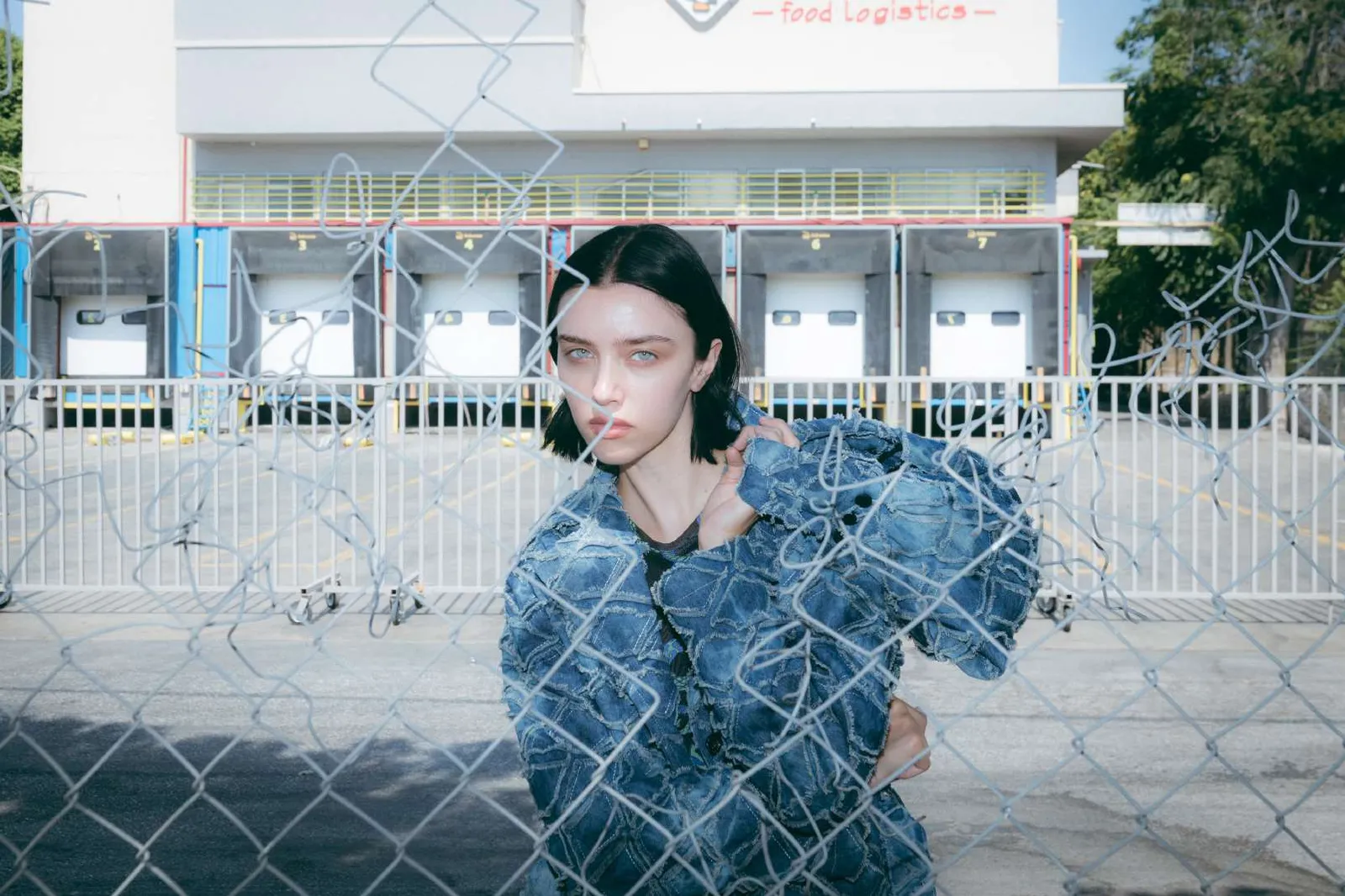 Model in a blue patchwork denim jacket behind a chain-link fence, a loading bay behind her