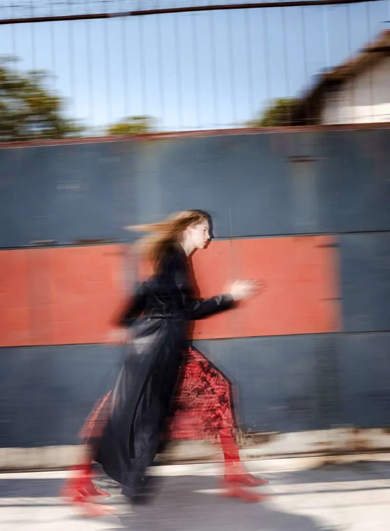 Model in a long black patent coat over a red and black printed dress and red boots, walking fast past a red and grey wall, blurred by motion