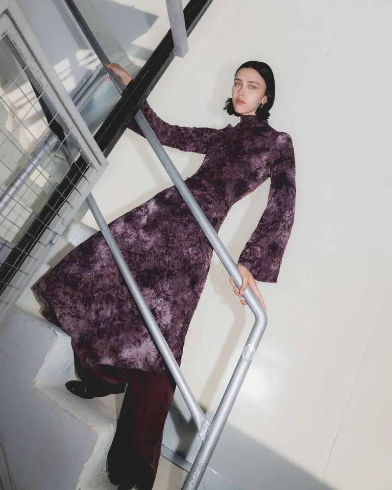 Model in a plum and black high-neck dress with flared sleeves over burgundy trousers, on a white staircase