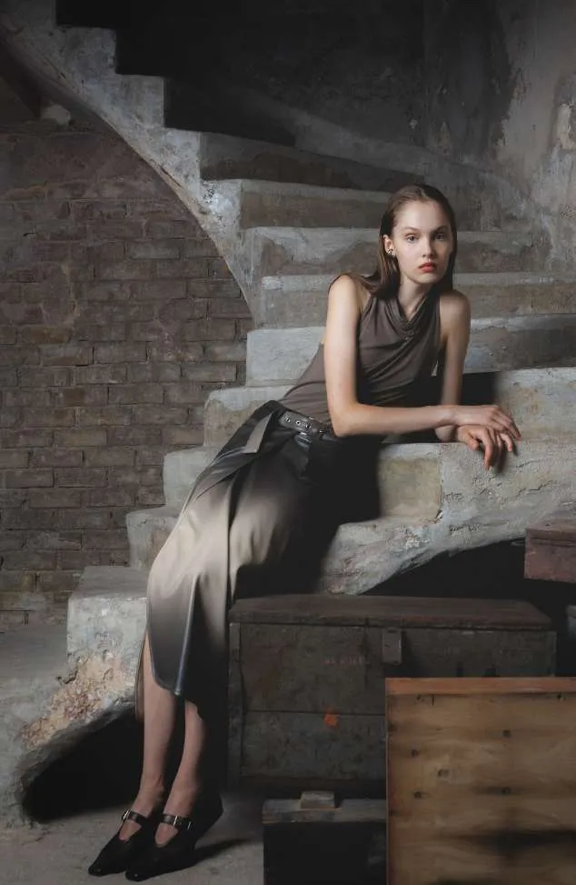 Model in a taupe draped sleeveless top and a satin maxi skirt, seated on a concrete spiral staircase