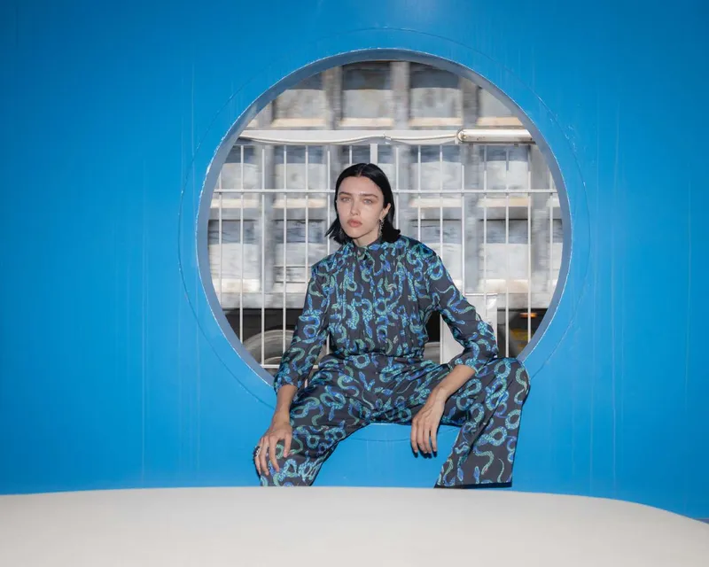 Model in a navy shirt and trousers with a blue and green snake-like print, crouched in a round window cut into a bright blue wall