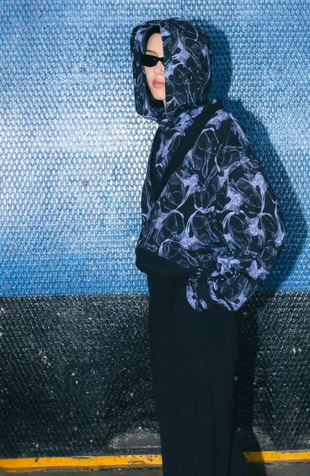 Model in a hooded jacket with a lilac and navy swirl print and black trousers, against a blue textured wall